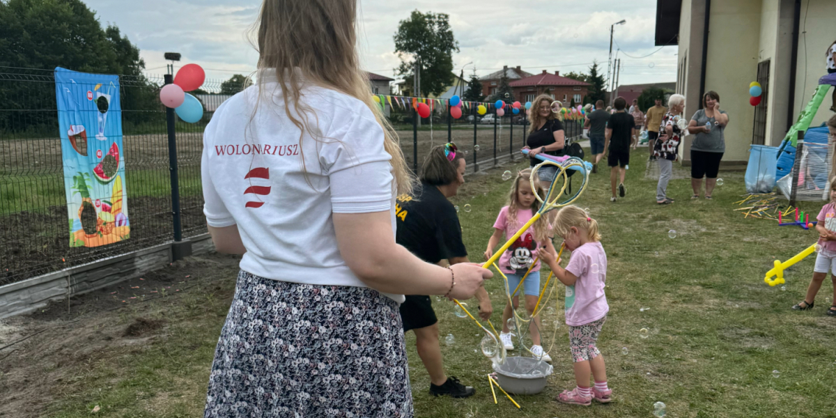 Młoda kobieta w koszulce z napisem Wolontariusz z logotypem PFR puszcza bańki wraz z dziećmi